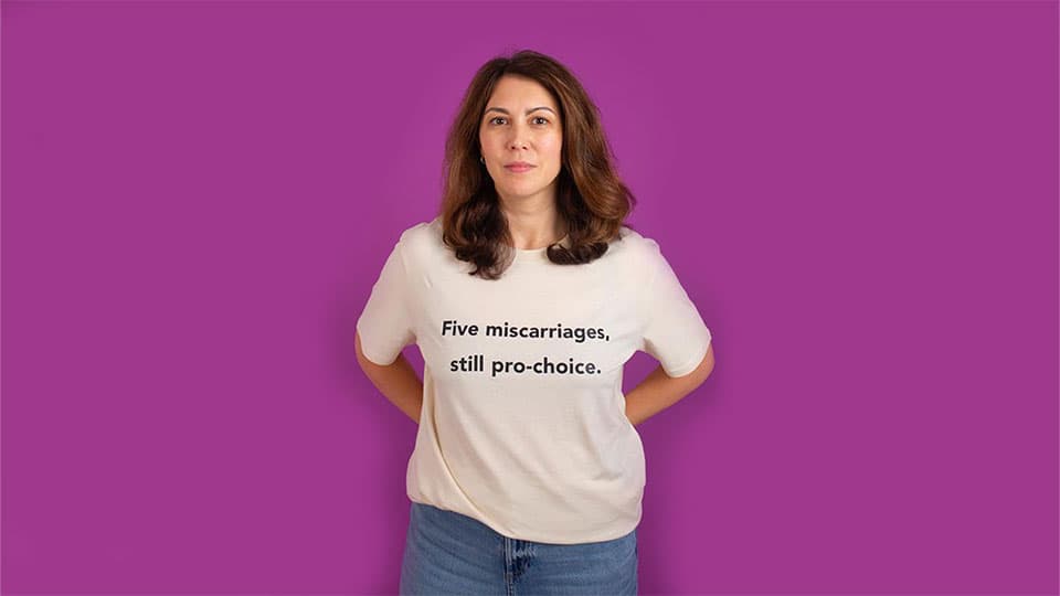 A woman wearing a t-shirt against a plain background which says five miscarriages, still pro-choice